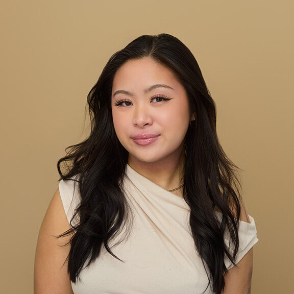 Portrait of a women with long, wavy, and black hair. She is looking and smirking at the camera while wearing a beige, high neck-slanted top.